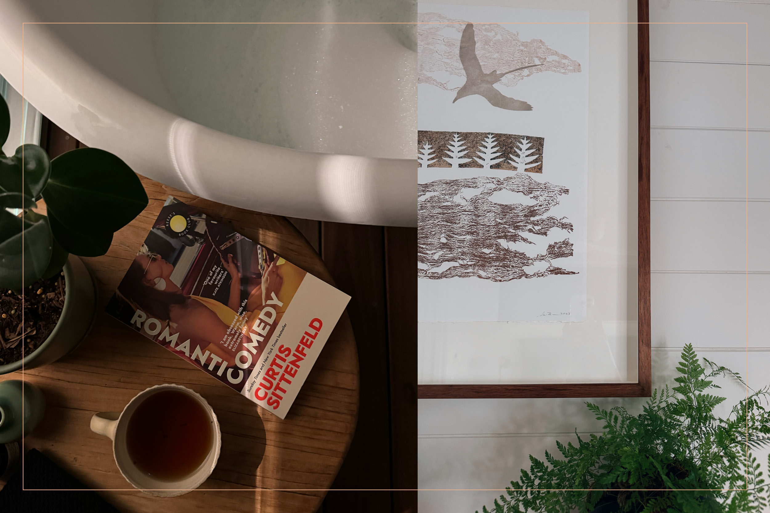 A photo of an outdoor bath with the book Romantic Comedy next to a cup of tea. Artwork on the wall with a fern plant underneath. 